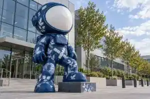 Three-quarter view of a 3.2 m Prussian-blue astronaut sculpture with soft white photographic shapes