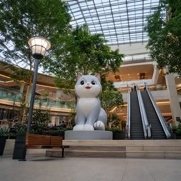Large cartoon-style fibreglass cat sculpture installed in a shopping mall atrium, with an escalator and seating for scale